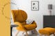Retro fauteuil van oranjegeel fluweel met bijpassende hocker, op een zwart-wit vloerkleed, plus een zwart bijzettafeltje, een matgrijs dressoir, een decoratieve spiegel, een betonnen tafellamp en een afbeelding van Audrey Hepburn; ernaast een zwarte metalen hanglamp.