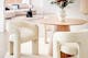 Runder Esstisch aus Holz mit Armlehnenstühlen aus beigem Boucléstoff, beides von der home24 Exklusivmarke Studio Copenhagen; dazu eine weiße, runde BUTLERS-Vase und eine Stehlampe aus Rattan. 