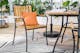 Gartenmöbel-Set im Industrial-Stil aus Akazienholz und schwarzem Stahl mit orangefarbenem Kissen auf einem Outdoor-Teppich mit schwarz-weißem Palmenprint.