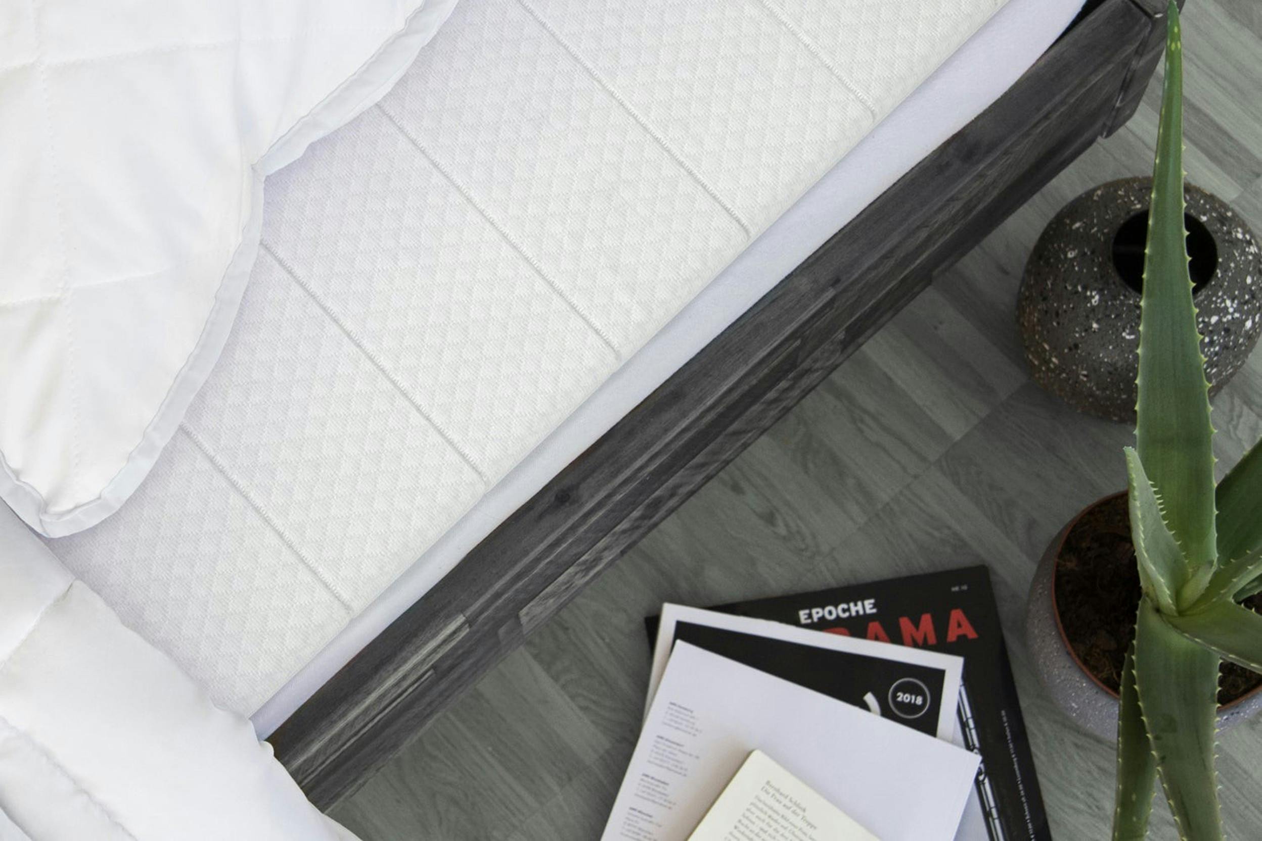 Draufsicht auf ein Bett mit Matratzen-Topper, daneben ein Kaktus und ein Zeitschriftenstapel