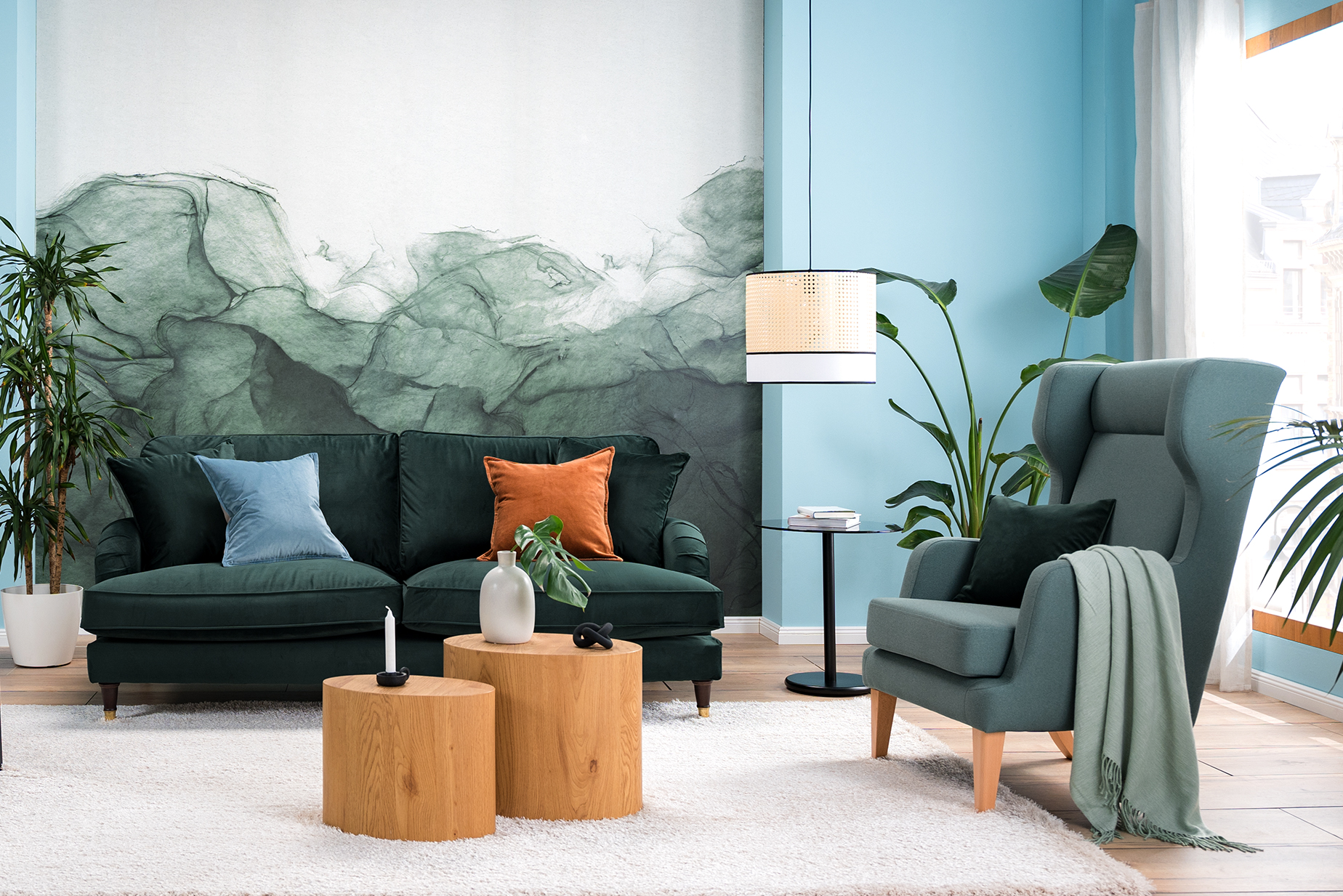 Wohnzimmer mit Samtsofa in Petrol, einem hellblauen Sessel und einem Retro-Sessel mit Wiener Geflecht, an der Wand eine blaugrüne Fototapete, dazu ein weißer Teppich, zwei Tische aus hellem Holz, ein schwarzer Beistelltisch, eine Pendelleuchte mit Wiener Geflecht sowie ein Sideboard, Deko und Wandbilder mit Tier- und Blumenmotiven.