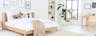Camera da letto con mobili in legno chiaro della serie Taylor di kollected, un marchio esclusivo di home24: un letto, un comodino, una sedia a dondolo e una scrivania con sedia in rattan, il tutto accostato a tessili color beige e marrone chiaro, tappeto, un pouf in macramè e piante.