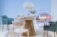 Runder Tisch mit hellen Holzsträngen, der von unterschiedlichen Stühlen im Skandi-Stil umgeben und mit Keramikgeschirr in zarten Pastelltönen sowie einem Trockenblumenstrauß gedeckt ist.