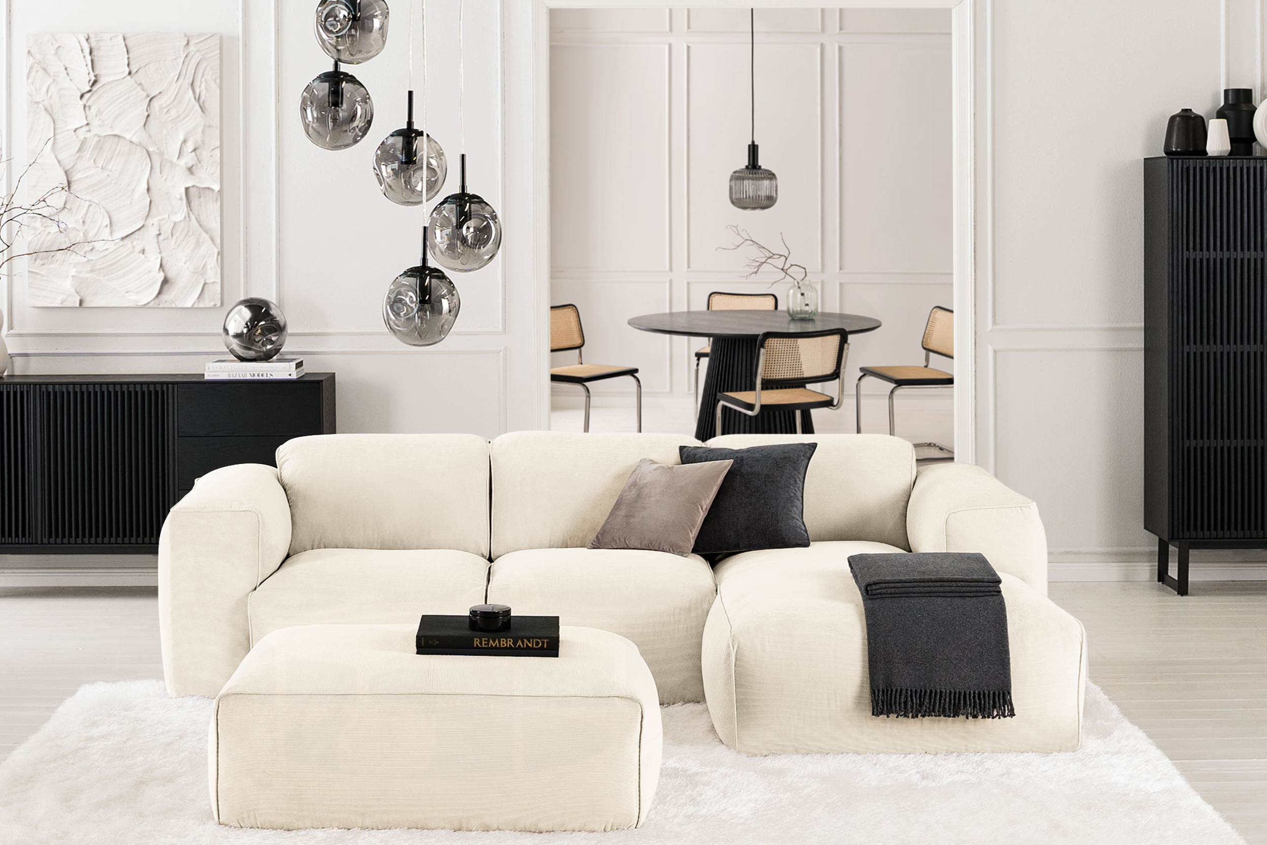 Wohnzimmer mit beigem Cordsofa und Cordhocker der Serie Hudson by Studio Copenhagen sowie Sideboard, Schrank und Esstisch der Serie Qara by Studio Copenhagen in Schwarz; dazu Pendelleuchten aus grauem Rauchglas, Flechtstühle und ein Servierwagen.