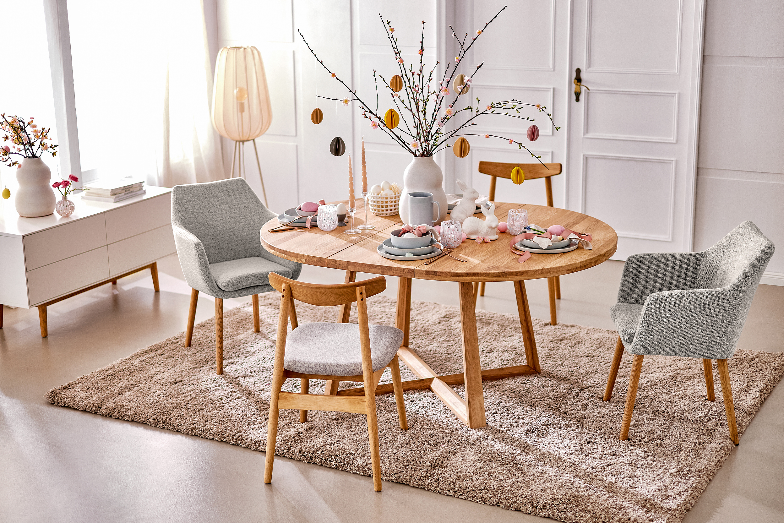 Esstisch Voksa der home24 Exklusivmarke Ars Natura, eingedeckt mit hellgrauem und weißem BUTLERS-Geschirr und bunter Osterdeko. Dazu passende Esszimmerstühle in Grau im Skandi-Stil, ein weißes Sideboard und ein beiger Teppich.