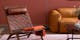 Wohnzimmer mit einem braunen Ledersessel in Flechtoptik, einem Ledersofa in Cognacbraun und einem schwarzen Couchtisch aus Glas und Metall. Dazu eine schwarze Stehleuchte, ein roséfarbener Teppich, ein Wandbild mit einer Hundezeichnung sowie eine orangefarbene Decke.