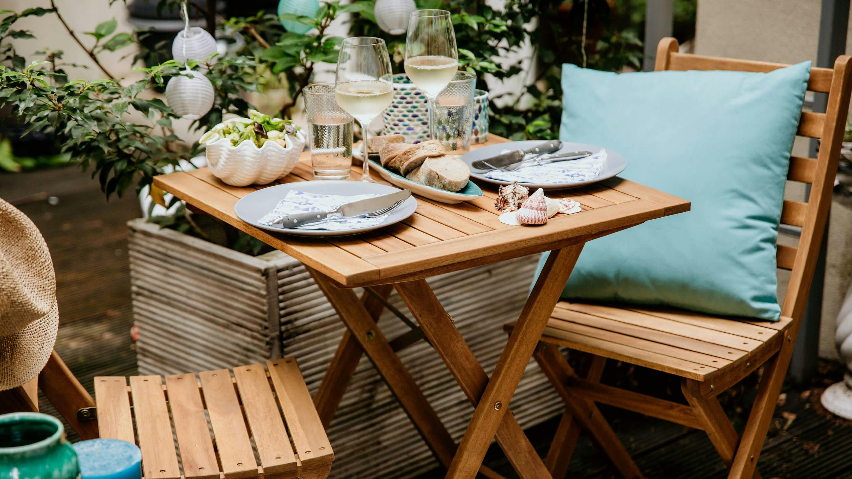 Gartentisch-Set aus Massivholz: 2 Klappstühle, 1 gedeckter Tisch mit u.a. gefüllten Weißweingläsern, etc. (Marke: Butlers).