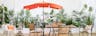 Garten im Boho-Stil mit Hängesessel, Windlichtern aus Bambus, einem schwarz-weißen Outdoor-Teppich und orangefarbenen Sonnenschirm, dazu Kissen und Plaids, viele Grünpflanzen sowie Gartenmöbel aus Akazienholz und Metall.
