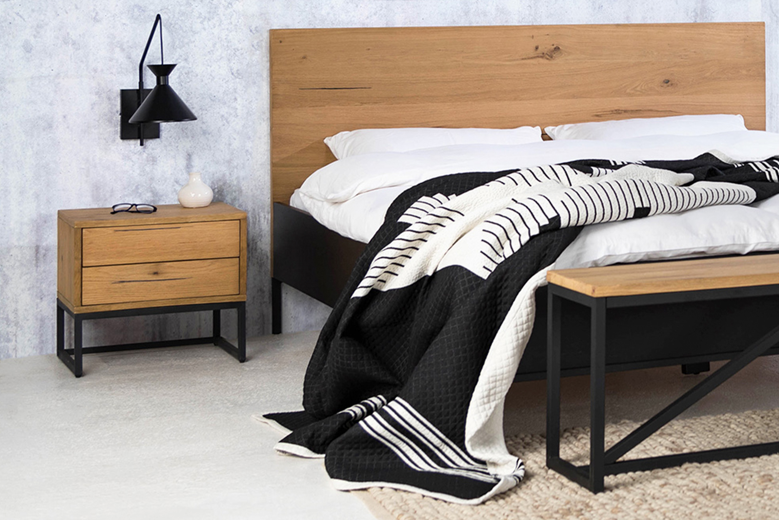 Schlafzimmer im minimalistischen Stil mit Bett, Nachttisch und Sitzbank der Serie Flox im Industrial-Stil aus Eichenholz und schwarzem Metall. Dazu schwarz-weiße Wandbilder, eine schwarze Wandleuchte, ein beiger Teppich und ein schwarzer Garderobenständer aus Metall.