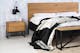 Slaapkamer in minimalistische stijl met bed, nachtkastje en bank van eikenhout en zwart metaal uit de industriële meubelserie Flox. Vergezeld van zwart-witte muurschilderingen, een zwarte wandlamp, een beige vloerkleed en een zwarte metalen kapstok.