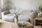 Soggiorno in stile scandinavo con divano Hudson bianco e tavolino da salotto rotondo in legno su tappeto a pelo corto color corda, in primo piano sulla destra, un divano panoramico grigio chiaro.