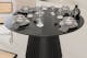 Table ronde noire avec piètement conique plissé, dressée avec de la vaisselle en céramique grise et des couverts et serviettes foncés.