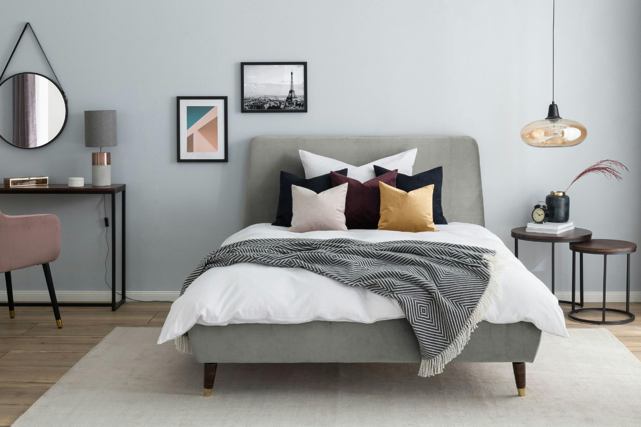 Chambre à coucher, avec lit gris clair, literie blanche et coussins colorés, tables de chevet en bois sombre rondes, cadres photos accrochés au mur, secrétaire en bois sombre, fauteuil rose poudré, miroir rond accroché au mur, quelques lampes