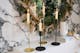Des bougies blanches dans des chandeliers dorés et noirs devant une couronne de Noël sur un mur en marbre ; à côté, des bougies colorées en spirale ainsi que des bougies lisses.