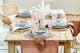 Mobili per la sala da pranzo in legno chiaro e rattan, stoviglie di ceramica, bicchieri rosa, posate color oro su runner di cotone.