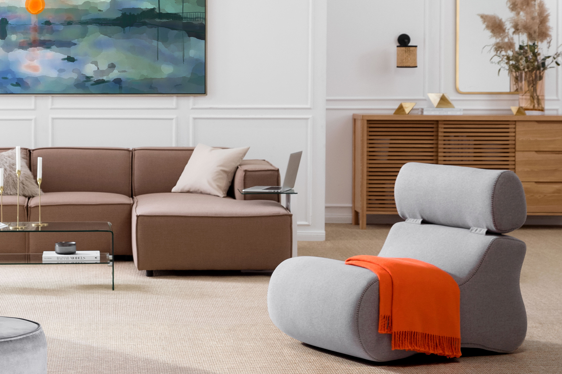 Wohnzimmer mit braunem Sofa, einem Sideboard aus Holz, Couchtisch aus Glas und einem grauen Sessel, dazu goldfarbene Dekoration.