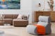 Wohnzimmer mit braunem Sofa, einem Sideboard aus Holz, Couchtisch aus Glas und einem grauen Sessel, dazu goldfarbene Dekoration.