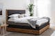 Lit boxspring gris, noir et bois dans une chambre aux mêmes tons, avec linge de lit blanc et déco sobre, et meubles en bois