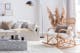Salon hygge avec un canapé blanc en tissu bouclette, une rocking chair en osier, une table basse en imitation marbre, des coussins et plaids douillets, des peaux de bête, un tapis shaggy couleur crème, un lampadaire noir, une enfilade en bois blond, des herbes de la pampa, des accessoires noirs et en rotin.