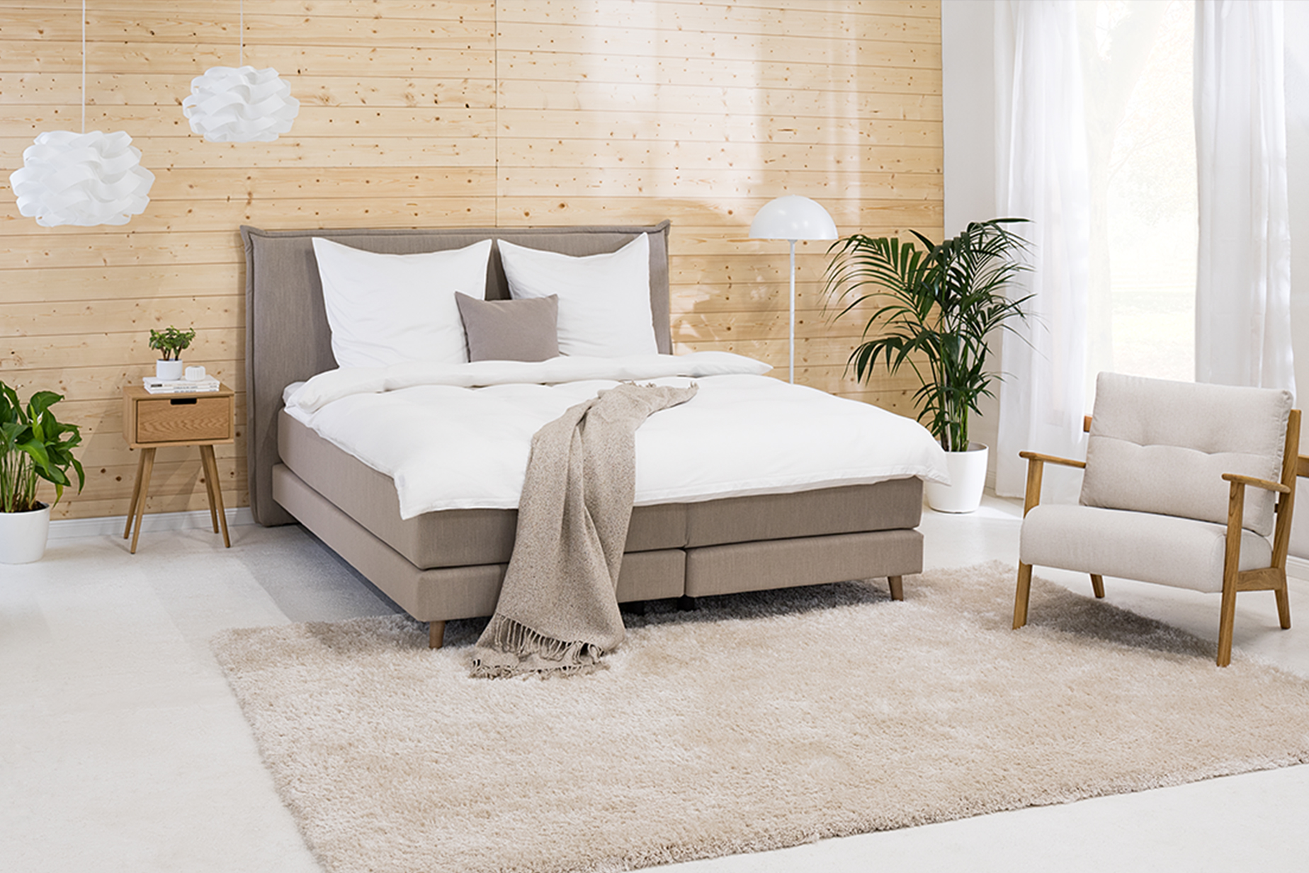 Schlafzimmer in Neutralfarben mit einem Bett, Kissen, Decke und Teppich in Taupetönen, dazu weiße Bettwäsche, ein weißes Sideboard, ein Nachttisch aus Holz, ein Sessel im Retro-Stil, Grünpflanzen, eine weiße Stehleuchte sowie weiße Pendelleuchten.