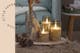 Arrangement aus drei LED-Kerzen unterschiedlicher Höhen sowie natürlicher Winterdeko auf einem Holzbrett; daneben ein Bild von einem Holzstuhl, auf dem zwei Adventskränze mit einer Lichterkette beleuchtet sind.
