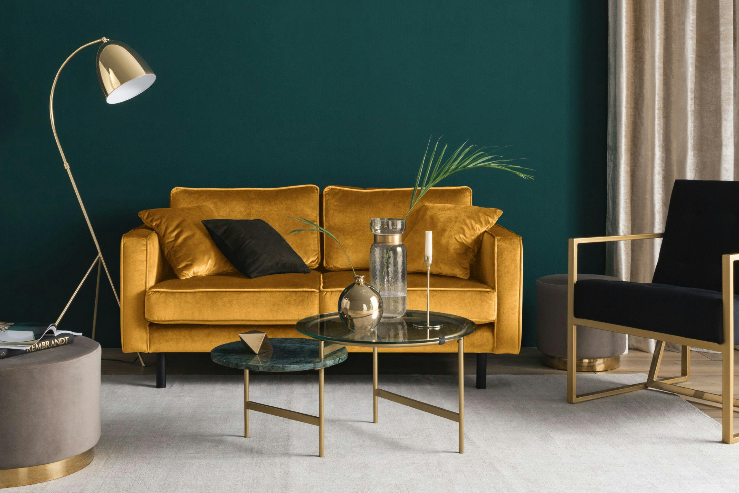 Gelbes Sofa aus edlem Samt vor tannengrün gestrichener Wand mit goldenen Accessoires