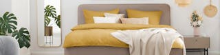 Beigefarbenes Bett mit gelber Bettwäsche, links ein Wandspiegel mit Grünpflanze davor, rechts neben dem Bett ein beigefarbener Nachttisch mit einer Blumenvase darauf
