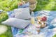 Gartenmöbelset aus Holz von BUTLERS, platziert auf einem grünen Rasen für ein Picknick: dazu Geschirr, Gläser und eine Etagere sowie ein weißes Kissen und eine blaue Tischdecke.