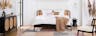 Camera da letto in stile rétro con letto davanti a una tenda bianca, poltrona e madia nere con inserti in paglia di Vienna, su tappeto moderno con motivo a quadri color pastello 
