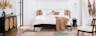 Slaapkamer in mid century modern met een bed, dressoir en fauteuil in donker hout en Weens vlechtwerk. Plus wit beddengoed, beige textiel, een zwarte hanglamp, vazen met pampagras, een vloerkleed met patroon in gedempte kleuren en een zwart nachtkastje.