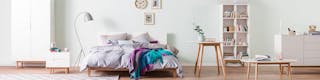 Bett mit grauer Bettwäsche und grauer Stehleuchte, weißer Schrank, Regal, Sideboard und Holztische vor zartgrüner Wand