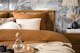 Slaapkamer in de boho-stijl met palmenbehang, meubels van het home24-merk kollected, bruin en wit textiel, een gebreide poef, vloerkleed, rotanlamp, zwarte spiegel en zwarte vazen.