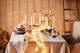 Winterlich gedeckter Tisch mit Leinentischdecke, schwarzem Geschirr, goldener Tischdeko und einer Lichterkette; im Hintergrund ist eine Holzvertäfelung und ein Holzstapel zu sehen