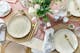 Bunt gedeckte Ostertafel mit beigem Geschirr, goldenem Besteck, geflochtenen Tischsets, frischen Blumen und Osterdeko.