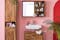 Badkamer met meubels uit de serie Deva by kollected, gemaakt van hoogwaardig sheeshamhout in de industriële stijl, tegen een roze muur. Het highboard staat voor een rode boog; daarnaast een binnenaanzicht van highboard Deva met veel opbergruimte voor badkamerspullen.