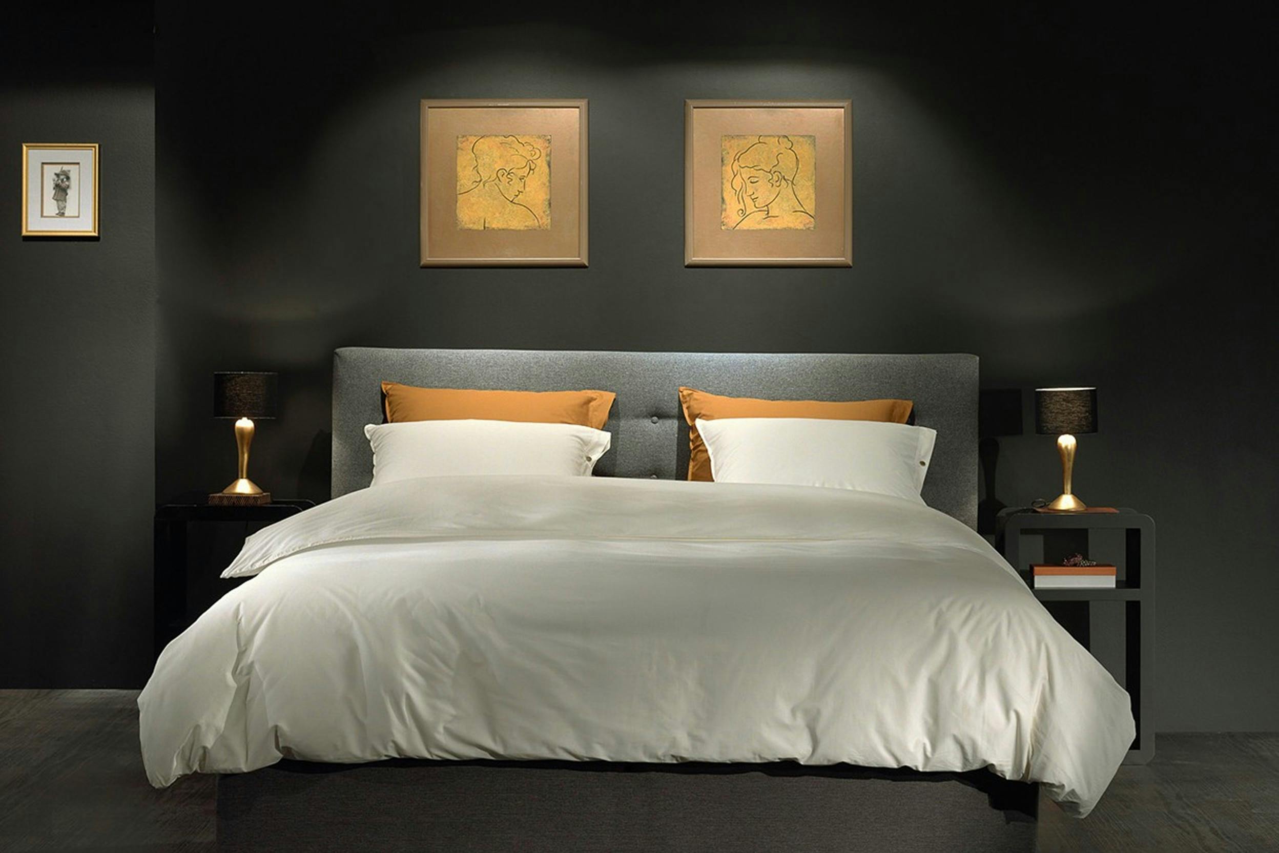 Grijs gestoffeerd bed met wit en geel beddengoed tegen zwarte achterwand met uitgelichte schilderijtjes, aan weerszijden nachtkastjes met schemerlampjes