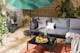 Outdoor-Loungesofa mit grauen Polstern, Kissen und Plaid, dazu ein schwarzer Tisch, Outdoor-Teppich und Grünpflanzen