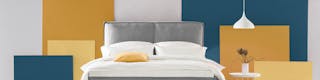 Photo d'un lit gris avec oreillers et draps blancs, contre un mur avec une table de chevet blanche. Le mur est jaune, blanc et bleu