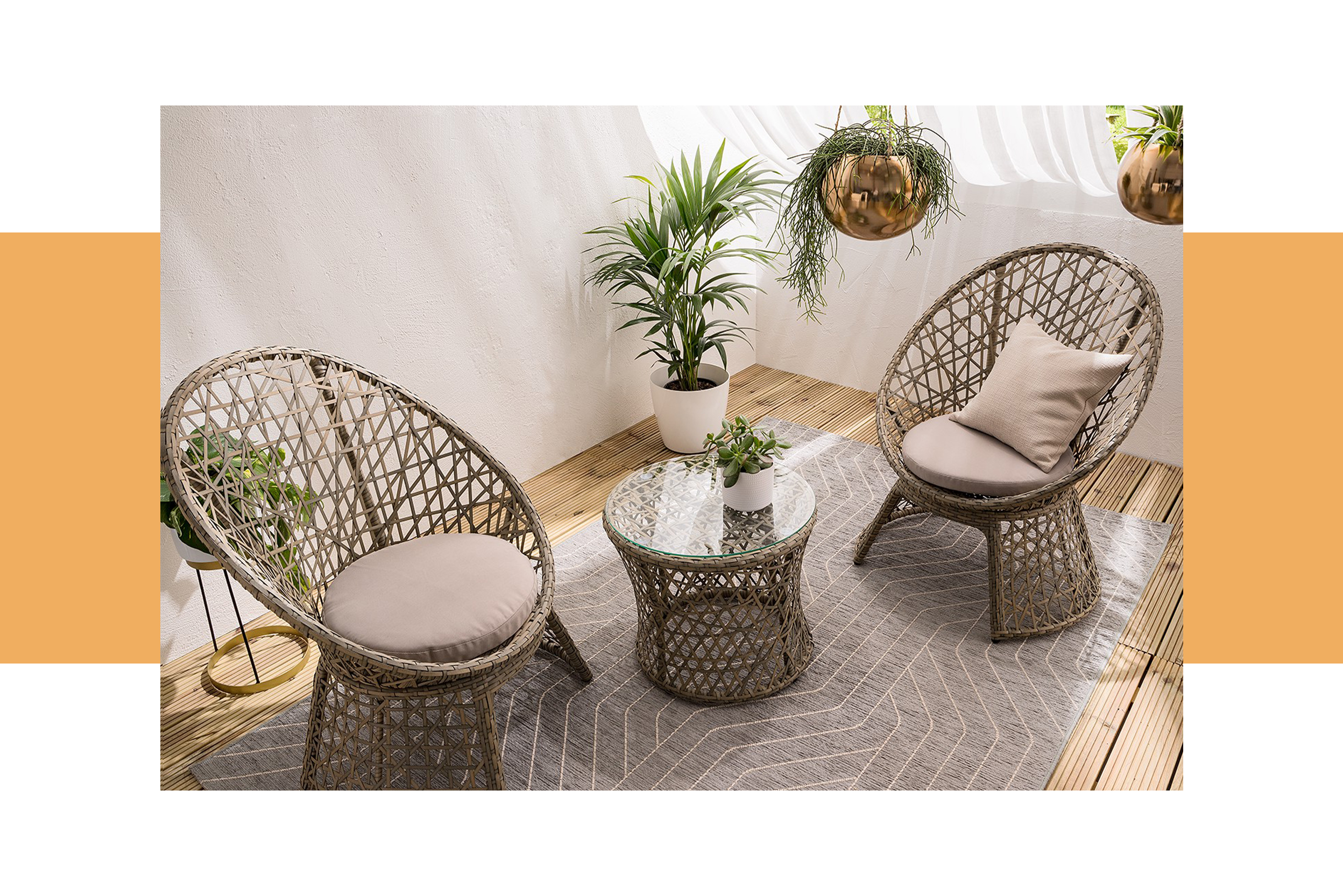 Outdoor-Rattanmöbel in einem Indoor-Wohnraum; zwei Rattansessel mit Kissen und ein runder Rattantisch mit Glasplatte, darunter ein grauer Outdoor-Teppich, ringsum viele Grünpflanzen wie Palmen und Sukkulenten