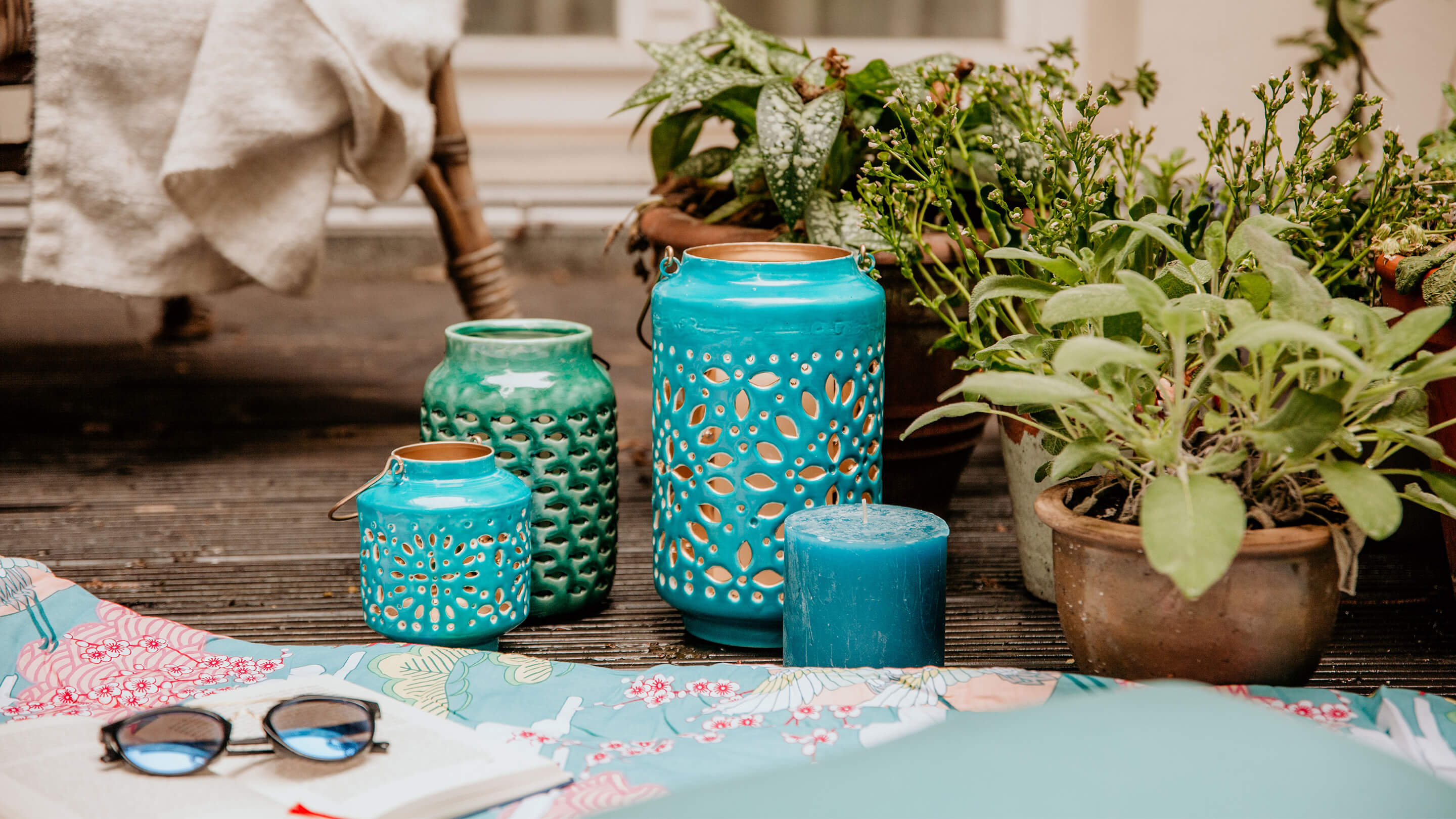 Windlichter in Türkis mit schönem Muster neben grünen Pflanzen, auf einem Terrassenboden (Marke: Butlers).