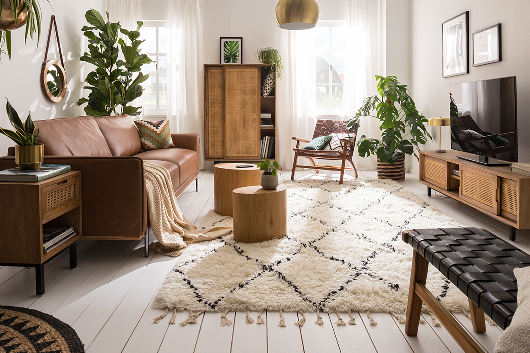 Wohnzimmer im Boho-Style mit einem Ledersofa, Holzmöbeln mit Wiener Geflecht, Rattan, vielen Pflanzen, Textilien und Deko aus Messing