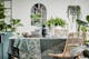 Eetkamer met houten tafel en stoelen van rotan omgeven door veel groene planten, plus schalen, serviesgoed in zwart en grijs en stijlvolle glazen van het merk BUTLERS.