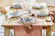 Gedekte brunchtafel, eetkamermeubels van natuurlijk hout en rotanweefsel, keramisch servies, roze glazen en goudkleurig bestek op een tafelloper van katoen.