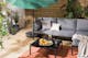 Outdoor-loungebank met grijze bekleding, kussen en plaid, daarnaast een zwarte tafel, outdoorvloerkleed en groene planten.