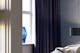 Blaue Tischlampe und grüne Stehleuchte in einem Raum, dahinter ein Schlafzimmer mit blauen Vorhängen, einer dunklen Pendelleuchte, grünem Bett mit Kissen und Decke