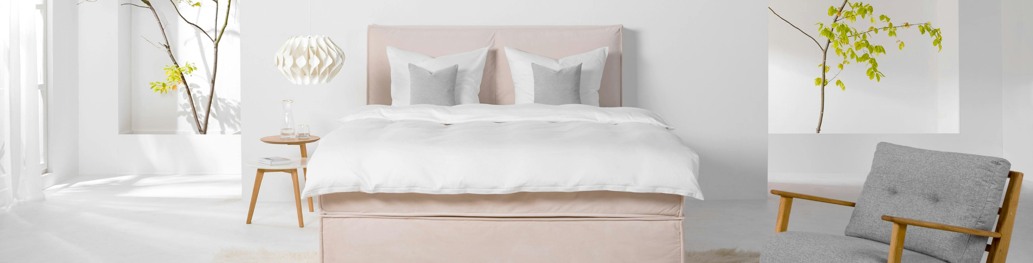 Rosa Boxspringbett mit weißer Bettwäsche in einem hellen Raum mit Sessel und Nachttisch