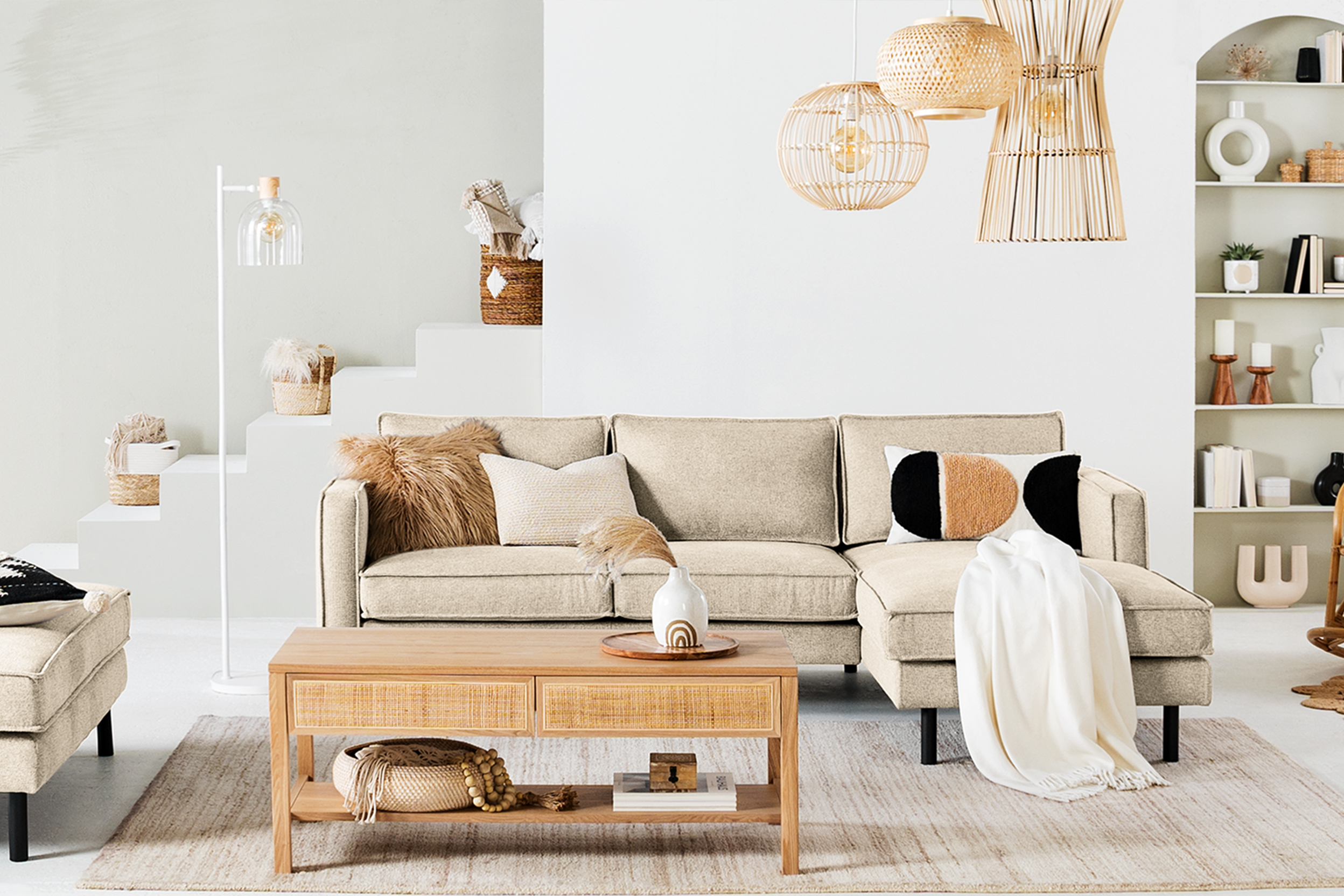 Wohnzimmer mit Möbeln der Serien Fort Dodge und Taylor der home24 Exklusivmarke kollected: Im Mittelpunkt ein beiges Sofa, dazu ein Couchtisch mit geflochtenen Applikationen, Pendelleuchten aus Rattan, Kissen, Teppich und Plaid in Beige- und Cremetönen sowie viele Dekoobjekte im Wandregal.