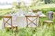 Festlich gedeckter Tisch auf einer grünen Wiese im Freien mit weisser Tischdecke, hellem Keramikgeschirr, Weingläsern und eleganter Tischdeko mit Blumen in weissen Vasen, dazu Landhausstühle aus Holz.