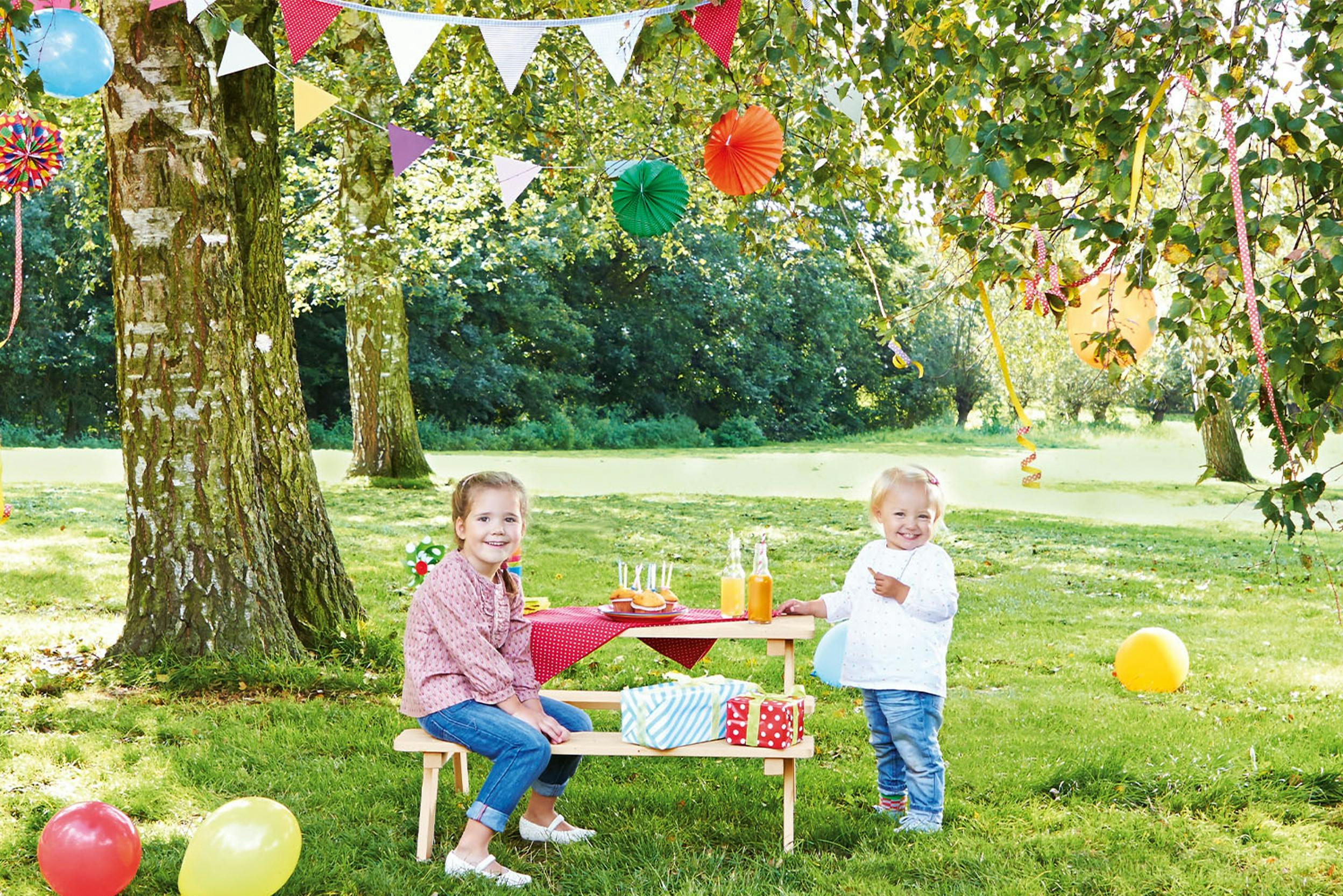 Kinder an einer Kindersitzgruppe unter einem Baum mit bunten Luftballons, Girlanden und Lampions