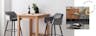 Table de bar en bois avec chaises de bar capitonnées de velours, desserte noire avec plante en pot et livres sur le plateau supérieur et bouteilles sur le plateau inférieur